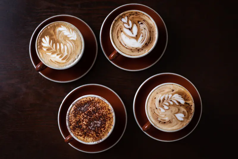 Four espresso drinks with milk seen from above on a dark wooden table, three with rosetta latte art and one dusted with chocolate powder.