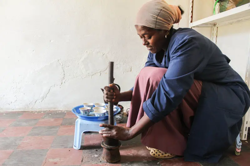 A woman crouches on a tiled floor pounding roasted coffee in a wooden mortar, beside a stool holding a tray of small handleless cups and a dark coffee pot.