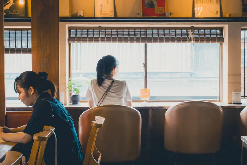 Two women sit in a wood-paneled café, one at a window counter with leather chairs and bamboo blinds, the other reading at a table.