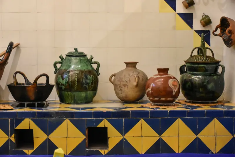 Green-glazed and painted clay pots stand on a blue and yellow tiled stove, with clay mugs hanging on the wall above.